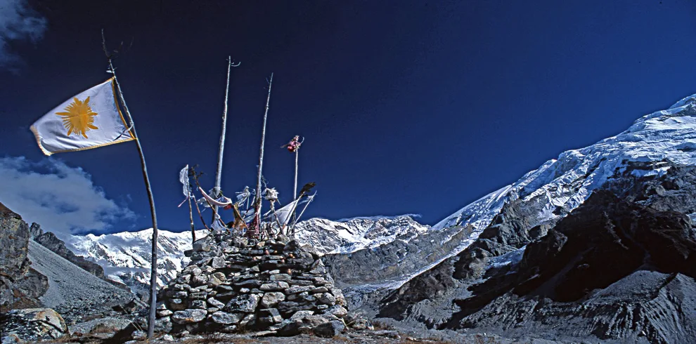 Kanchenjunga South Basecamp Trek 1 Prayer flags on a stone cairn with snow-covered kanchenjunga mountains under a deep blue himalayan sky in kanchenjunga south basecamp trek