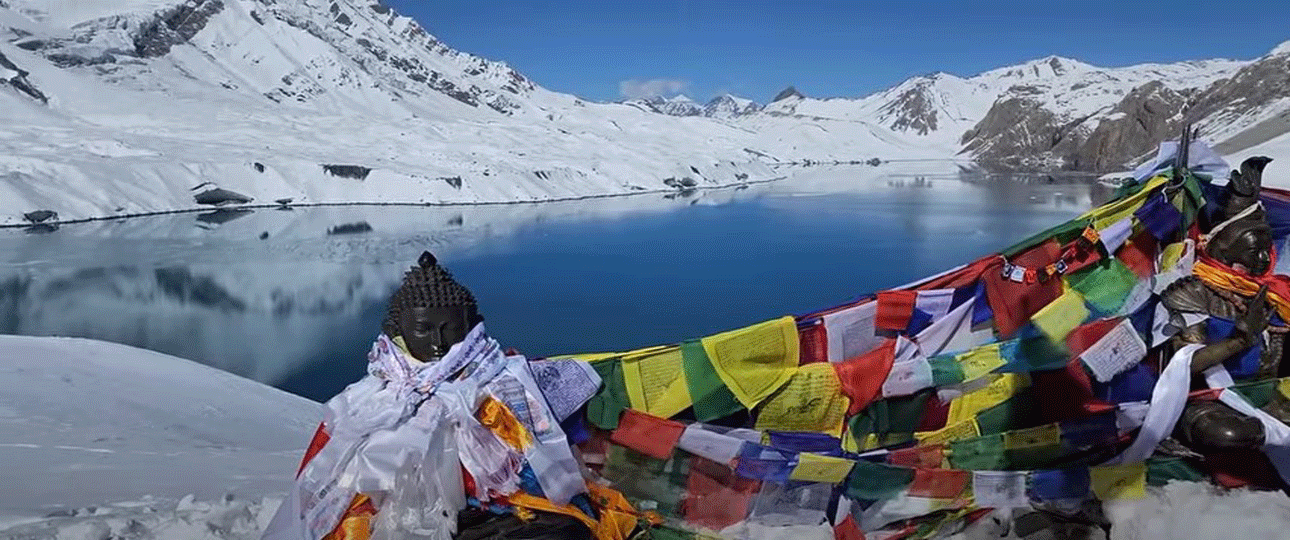 Frozen-blue tilicho lake surrounded by snow-covered himalayan peaks with prayer flags in the foreground.